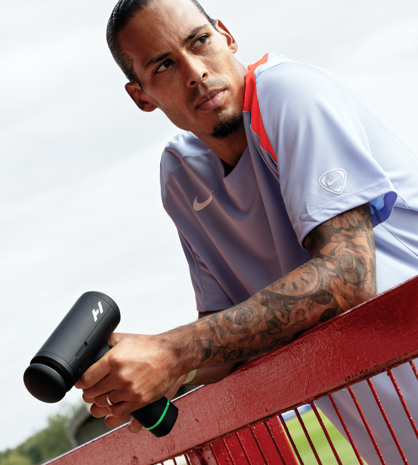 Virgil van Dijk holding a Hypervolt 3 Pro, outdoors, leaning on a red fence.