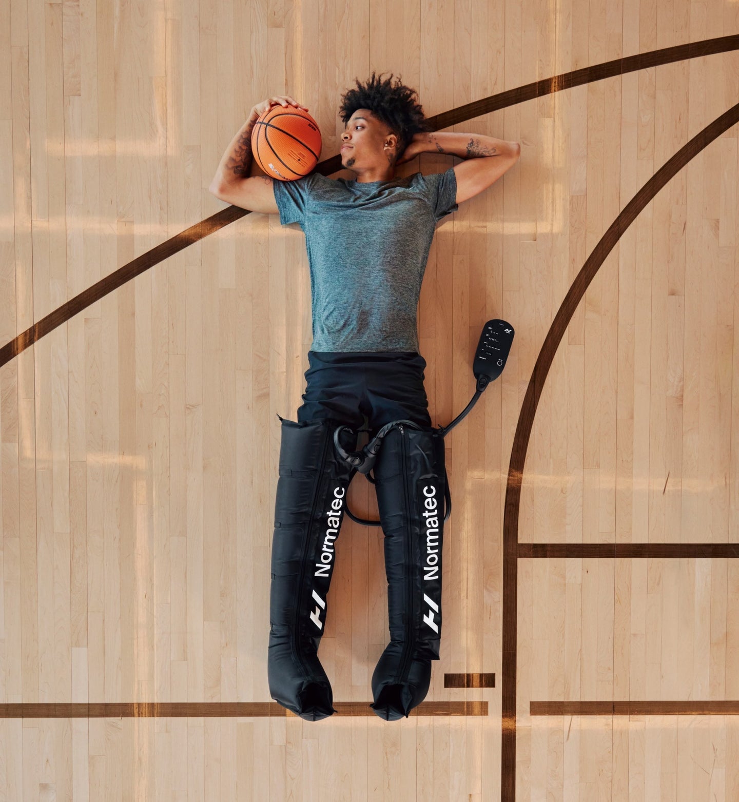Person lying on a basketball court holding a ball, wearing Normatec 3 legs compression sleeves